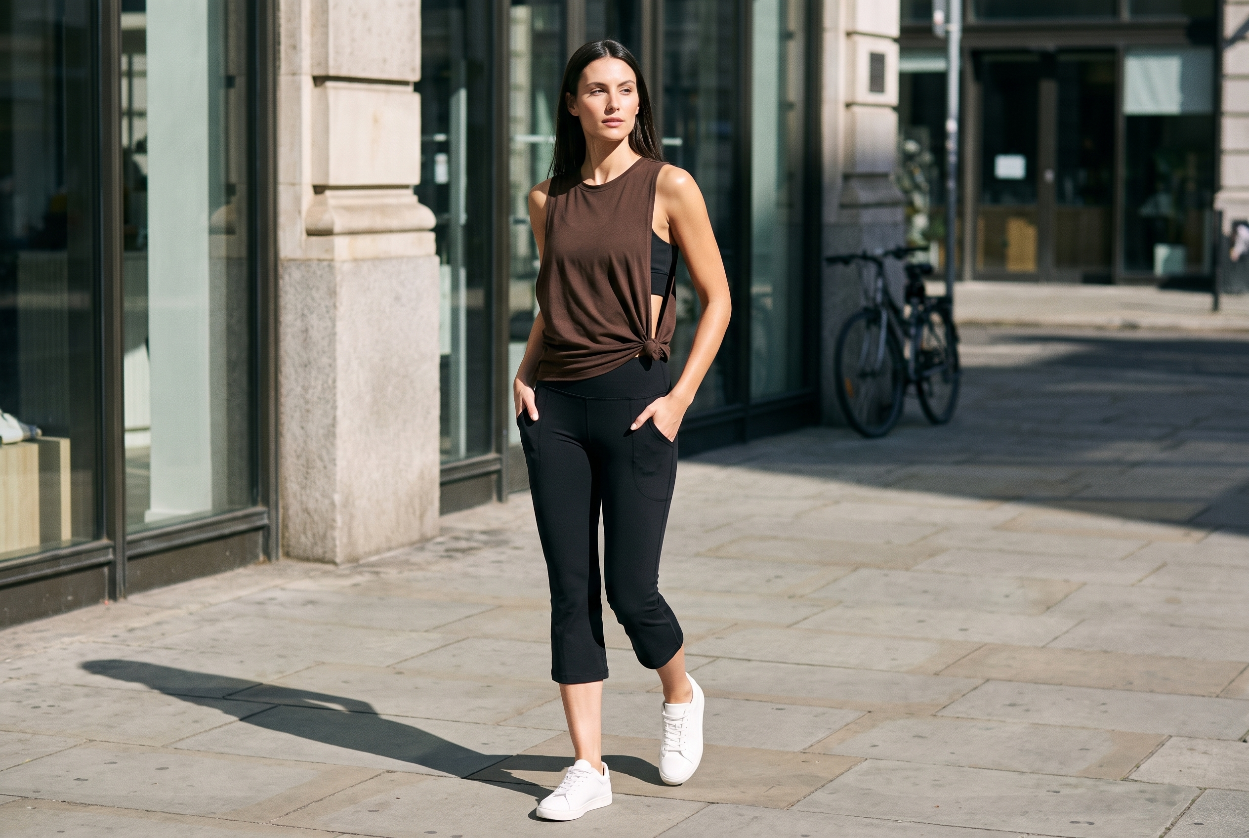 Woman walking on a city pavement with both hands in the deep thigh pockets of RYZ Core Active Flair Capri Leggings in black, worn with the Core Layer Tank in chocolate brown